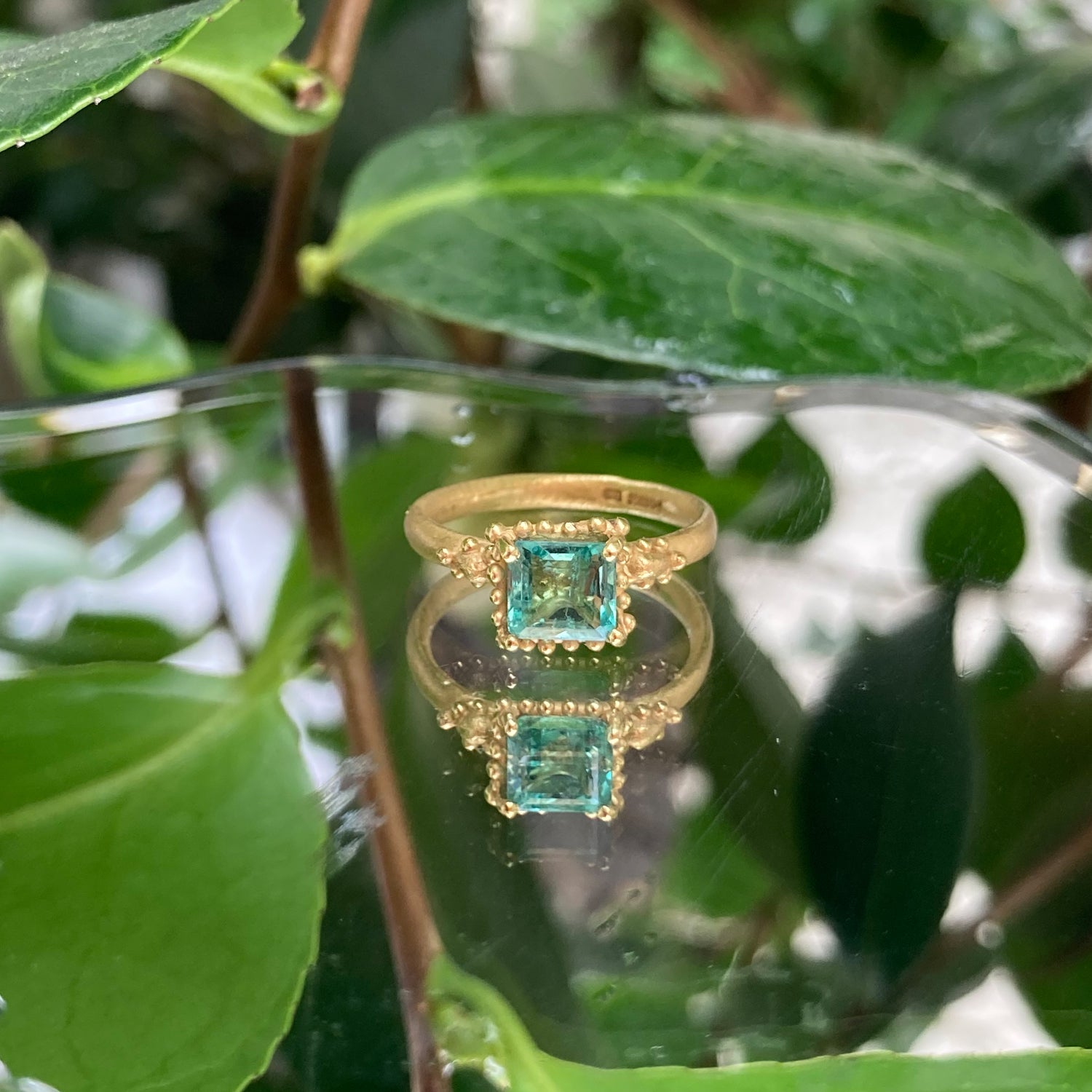 Ruth Tomlinson 18ct yellow gold ring with square facetted solitaire emerald with beaded granulation surround engagement ring. Alternative engagement ring on leaf background. Organic emerald ring.  